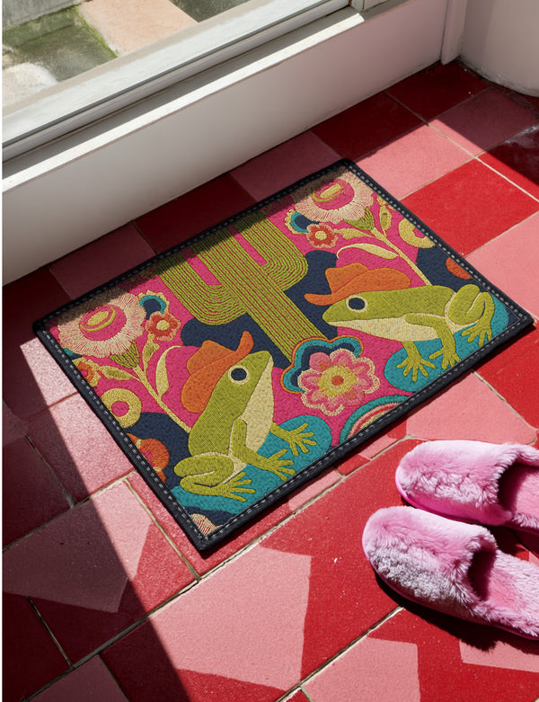 A rectangular polyester doormat with a black trim and non-slip rubber backing. The vibrant, colorful design features a symmetrical illustration on a dark navy blue background. In the center, two green frogs wearing orange cowboy hats sit facing each other. Between them is a tall, olive-green saguaro cactus. The scene is filled with large, psychedelic-style flowers in hot pink, orange, and yellow. The overall aesthetic is funky, retro 70s, maximalist, and trippy, mimicking the look of embroidery.